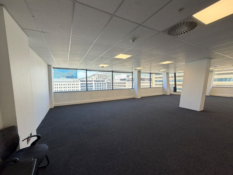 Spacious, open-plan office area featuring large windows with city views, suspended ceilings, and neutral carpeting, offering a bright and professional workspace.