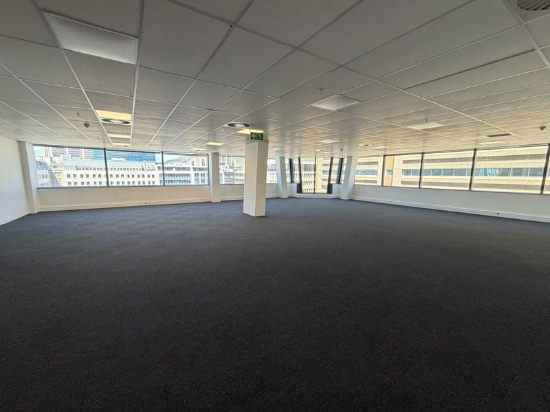 A spacious, open-plan office area featuring large windows with city views, suspended ceilings, and dark carpet tiles, offering a versatile workspace.