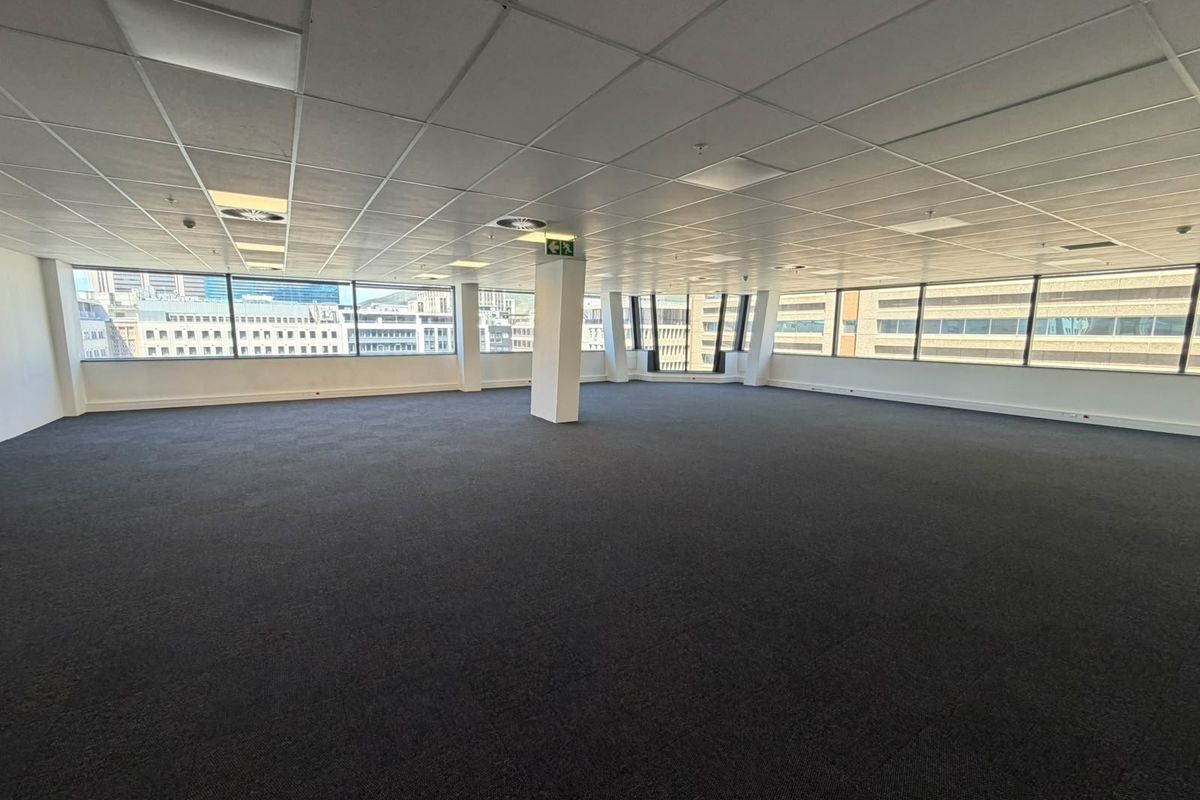 A spacious, open-plan office area featuring large windows with city views, suspended ceilings, and dark carpet tiles, offering a versatile workspace.