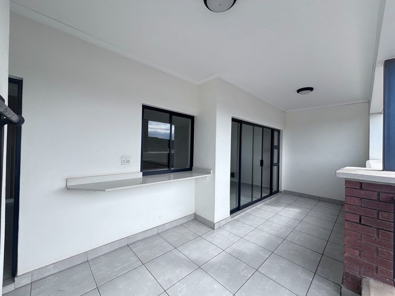 A clean, covered balcony featuring tiled flooring, a built-in stone serving counter, and sliding glass door access to the interior living space.