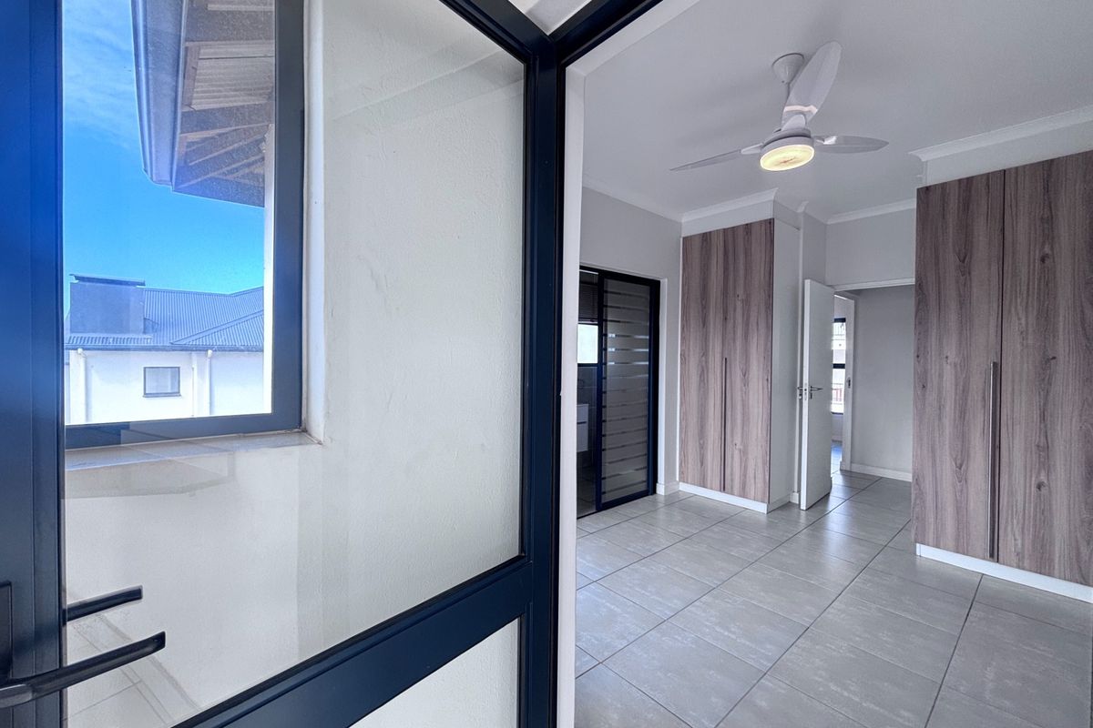 A bright, modern bedroom featuring neutral tiled flooring, built-in wood-finish cupboards, and a contemporary ceiling fan.