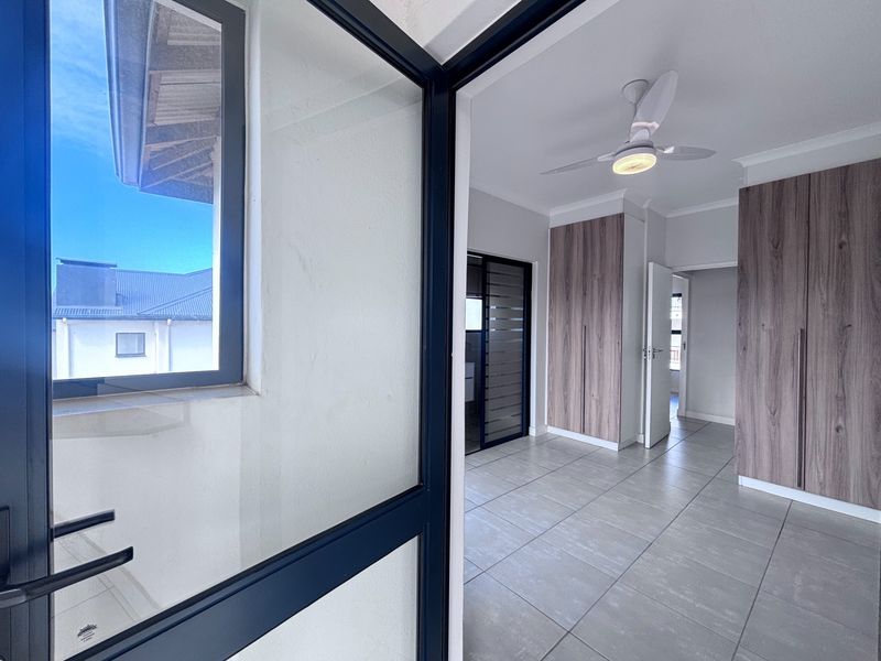 A bright, modern bedroom featuring neutral tiled flooring, built-in wood-finish cupboards, and a contemporary ceiling fan.