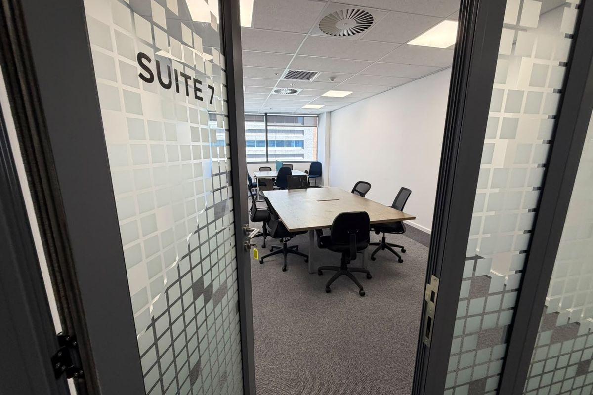 A professional office suite featuring a suspended ceiling, recessed lighting, air conditioning, and carpeted flooring, viewed through a glass entrance door.