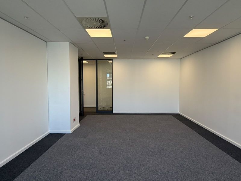 A clean, empty office space featuring a suspended ceiling with recessed lighting, air conditioning vents, and neutral carpet flooring with a glass partition door.