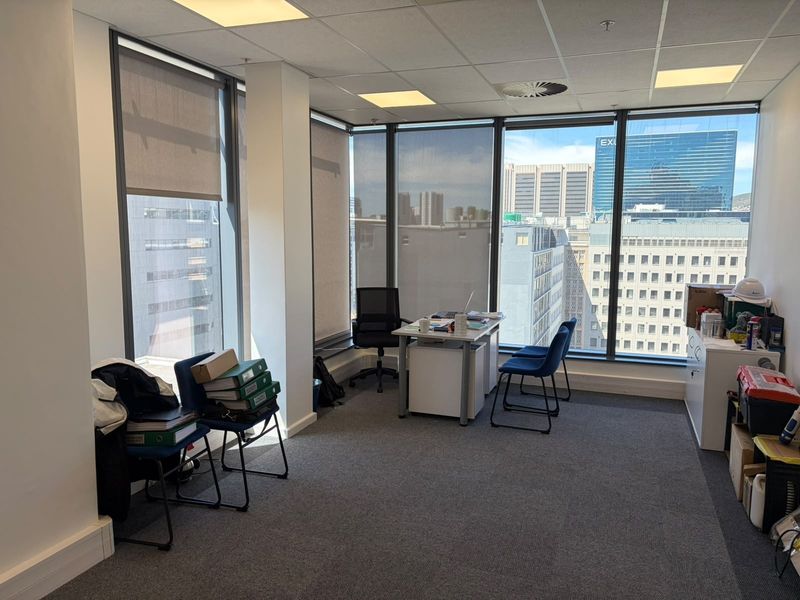 A bright office space featuring floor-to-ceiling windows with city views, carpeted flooring, and a suspended ceiling with integrated lighting.