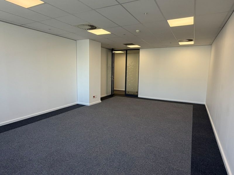 A clean, vacant office space featuring a suspended ceiling with recessed lighting, grey carpet flooring, and a modern glass partition door.