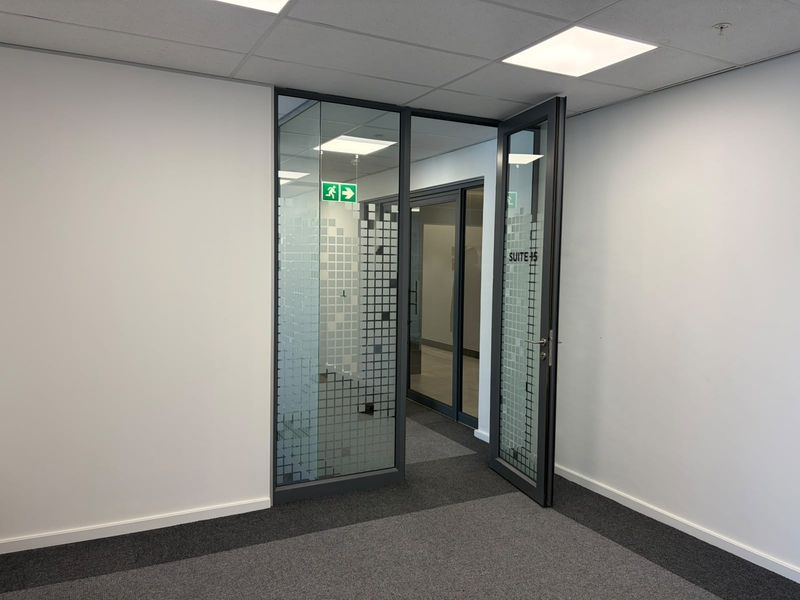 A modern office suite entrance featuring a glass partition wall, framed glass door, carpeted flooring, and recessed ceiling lighting.