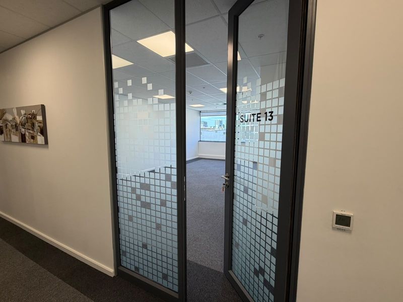 A bright, vacant office suite featuring glass entry doors, carpeted flooring, and a suspended ceiling with recessed lighting, offering a clean professional workspace.