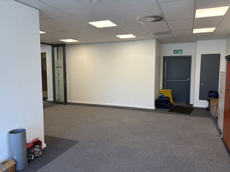 A clean, open-plan office space featuring neutral carpeted flooring, a suspended ceiling with recessed lighting, and a secure fire exit door.