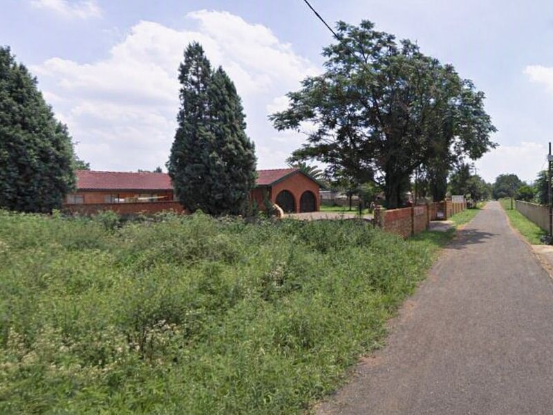 A street-level view of the residential property, featuring a paved road, brick boundary wall, and mature trees on the plot.