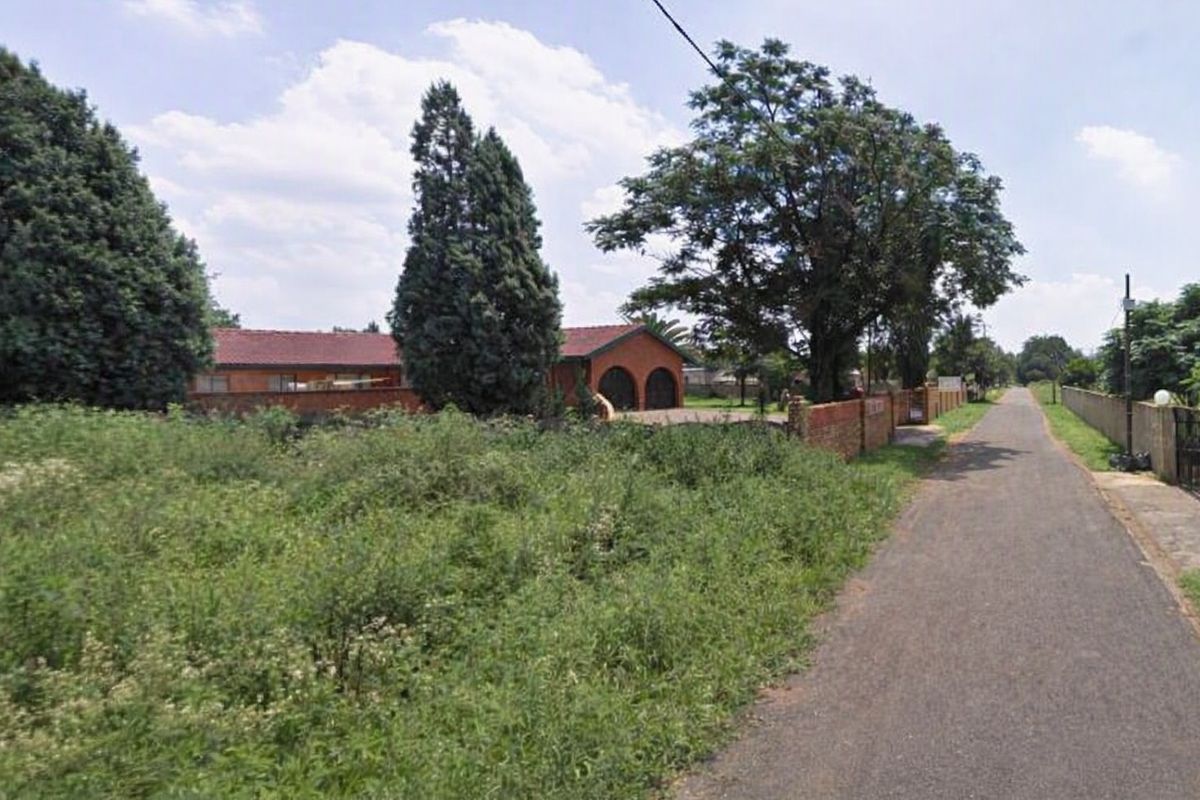 A street-level view of the residential property, featuring a paved road, brick boundary wall, and mature trees on the plot.