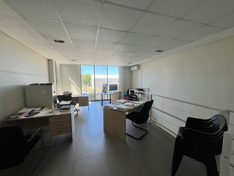 A functional office space featuring tiled flooring, a suspended ceiling, and a large window providing natural light, equipped with a wall-mounted air conditioning unit.