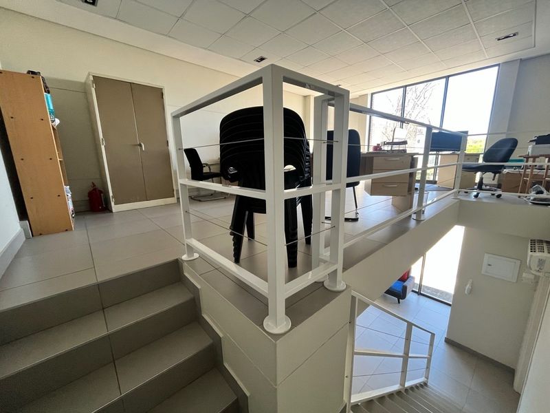 A split-level office space featuring tiled flooring, a modern metal balustrade, suspended ceiling, and large windows providing ample natural light.
