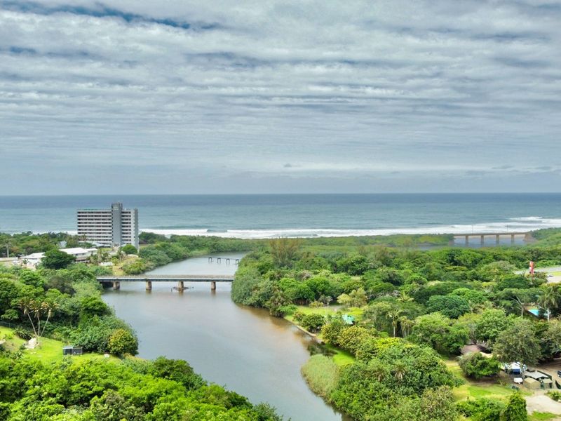 An aerial view of the coastal landscape featuring a river, bridge, and surrounding greenery with the ocean in the background.