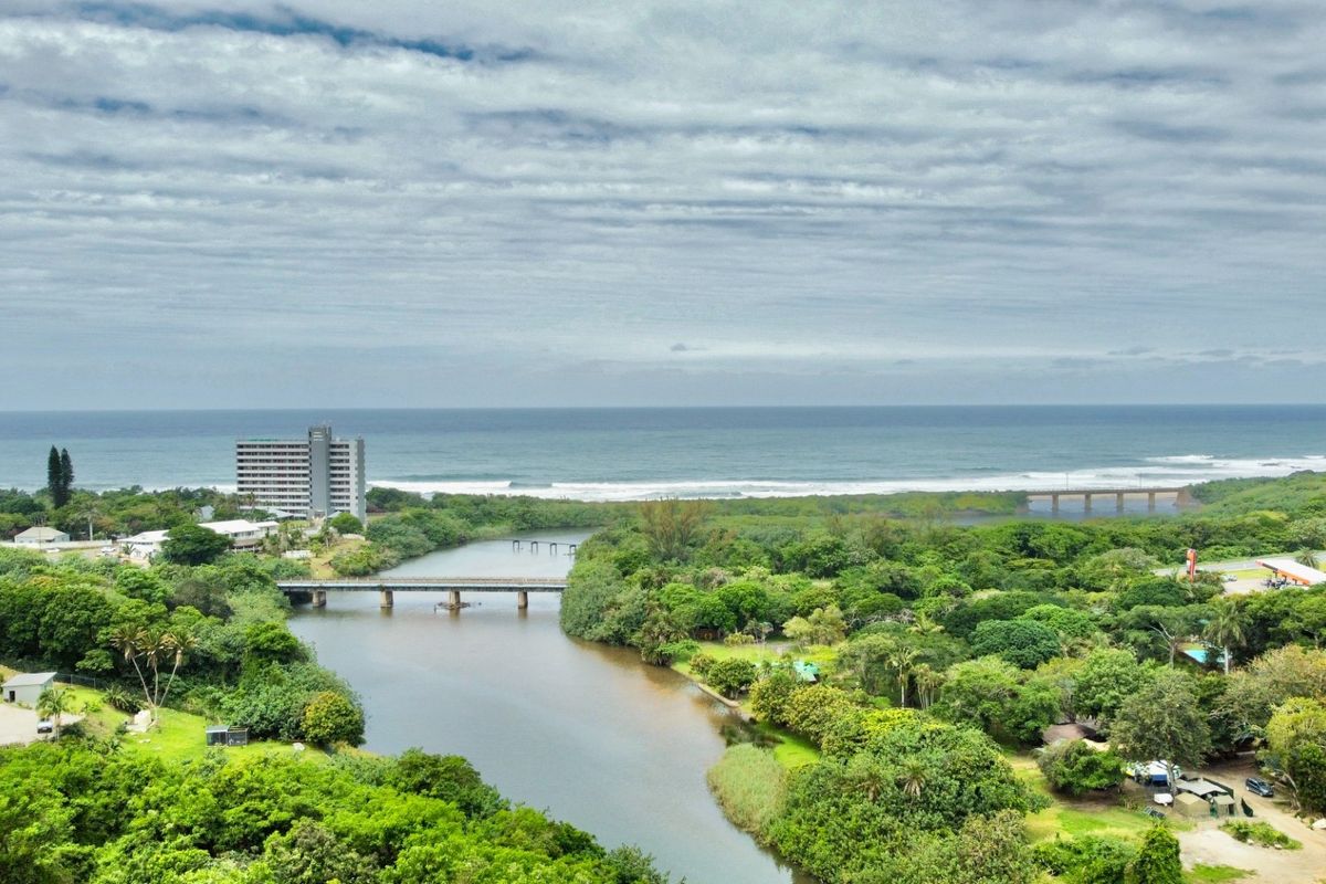 An aerial view of the coastal landscape featuring a river, bridge, and surrounding greenery with the ocean in the background.
