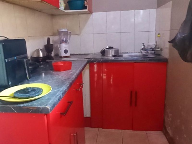 A compact kitchen featuring red cabinetry, a laminate countertop, and a stainless steel sink with tiled backsplash and flooring.