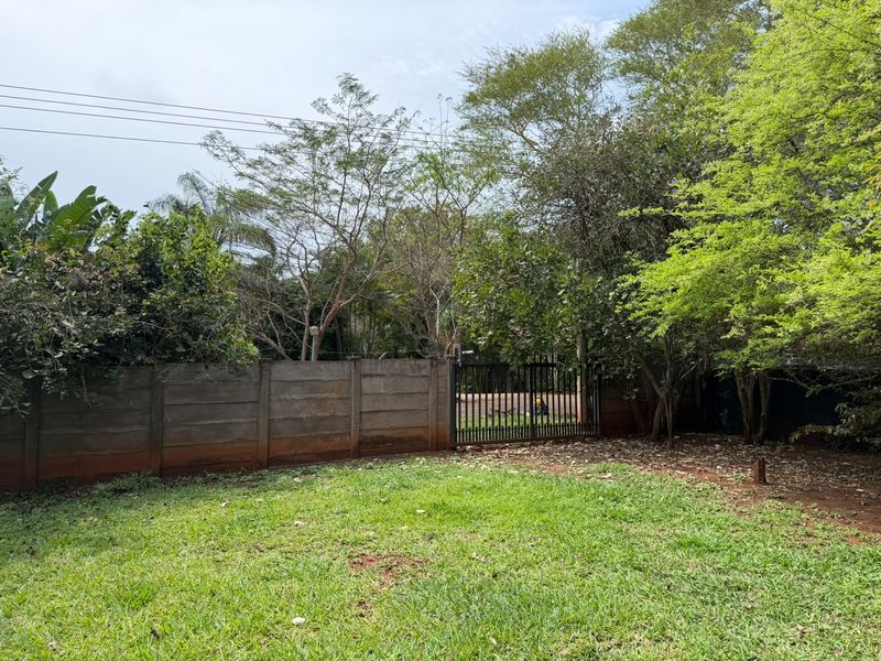 A spacious garden area featuring a lawn, mature trees, and a secure precast concrete boundary wall with a metal gate.