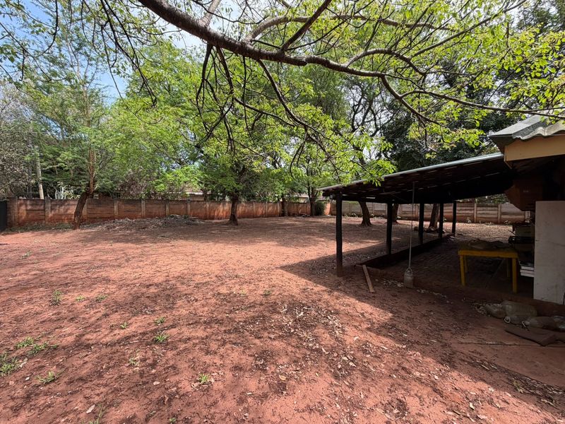 Spacious backyard featuring a large open area, mature trees, a brick boundary wall, and a covered patio structure.