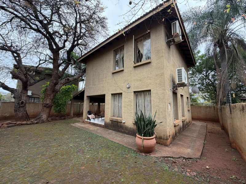 Rear view of a two-story house featuring a stucco facade, tiled roof, and a paved patio area set within a garden.