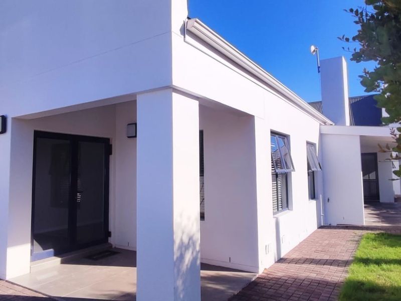 A well-maintained rear exterior view featuring a covered patio, paved walkway, and modern white facade with aluminium window frames.