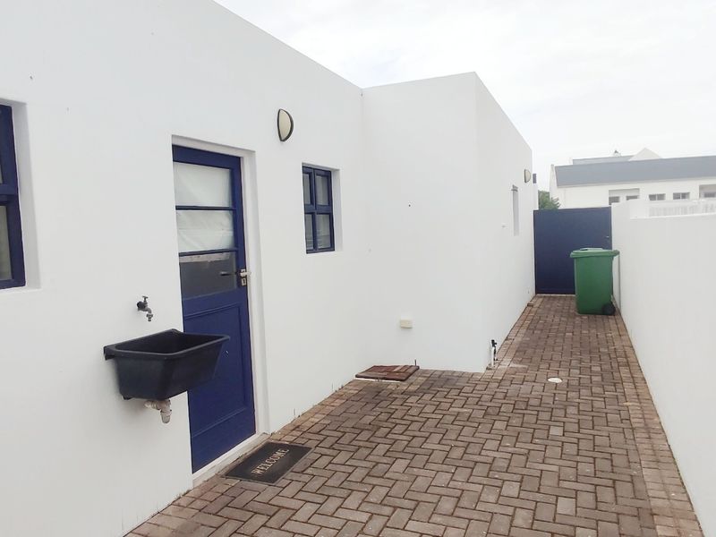 A clean rear exterior area featuring a paved walkway, white rendered walls, a blue external door, and a functional utility sink.