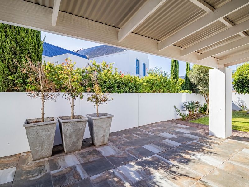 A covered patio area featuring tiled flooring, a corrugated roof structure, and a clean boundary wall overlooking a garden.