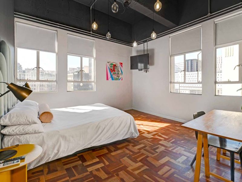 Spacious bedroom featuring warm parquet flooring, high exposed ceilings with industrial lighting, and large windows providing ample natural light and city views.