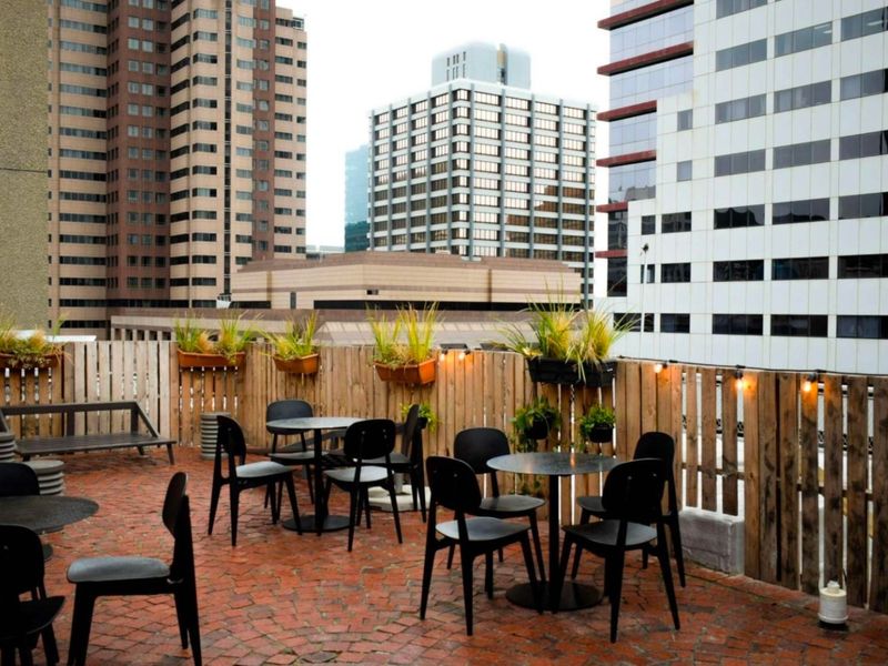 An urban balcony terrace featuring brick paving, timber slat fencing, and string lighting, offering a view of surrounding city high-rise buildings.