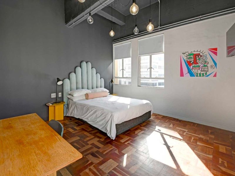 Spacious bedroom featuring warm parquet flooring, exposed industrial-style ceiling beams, and large windows with roller blinds, creating a bright and modern living space.