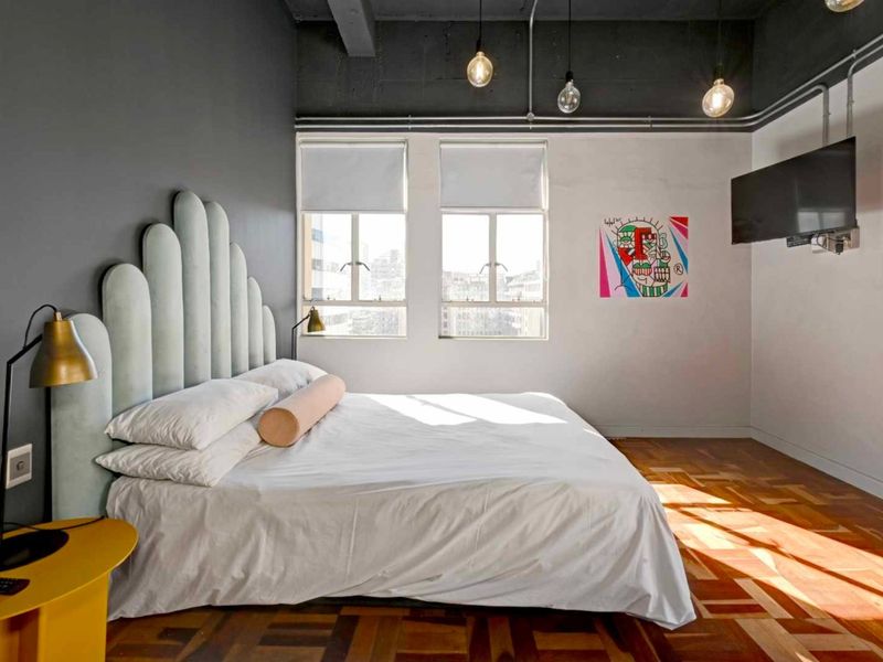 A modern bedroom featuring industrial-style exposed ceilings, parquet flooring, and large windows that provide ample natural light.