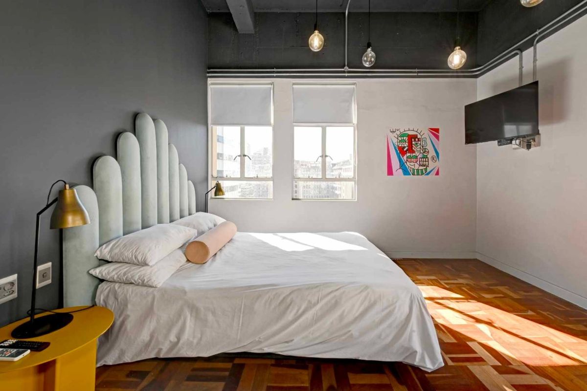 A modern bedroom featuring industrial-style exposed ceilings, parquet flooring, and large windows that provide ample natural light.