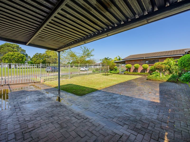 A covered outdoor patio area featuring paved flooring, a corrugated metal roof, and views of the front garden and security fencing.