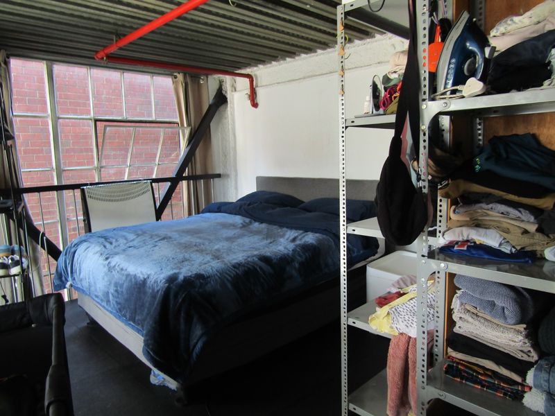Industrial-style bedroom featuring exposed corrugated metal ceilings, red fire sprinkler piping, and large steel-framed windows providing natural light.