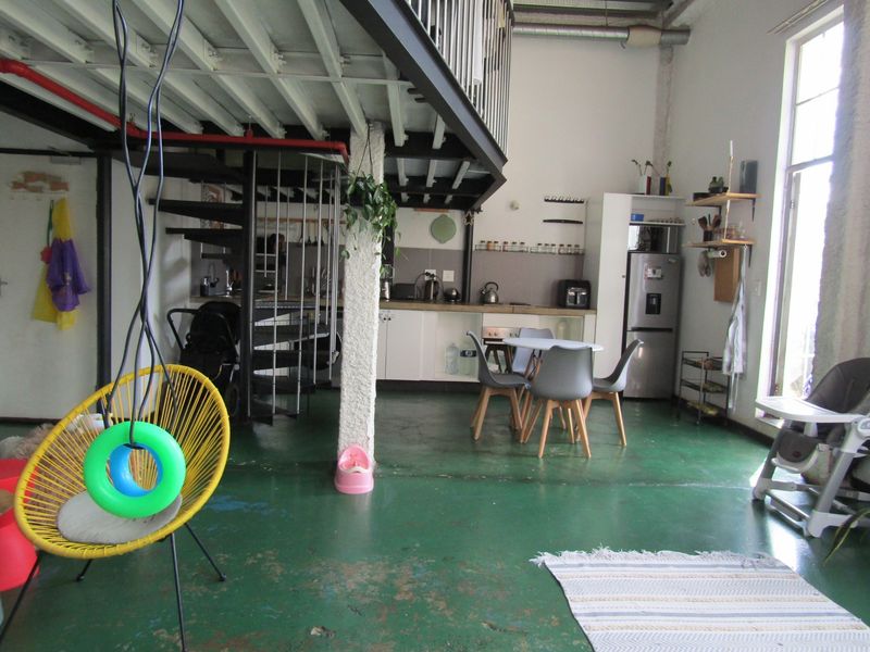 Open-plan living area featuring a mezzanine level with a spiral staircase, industrial-style windows, and a functional kitchen space with painted concrete flooring.