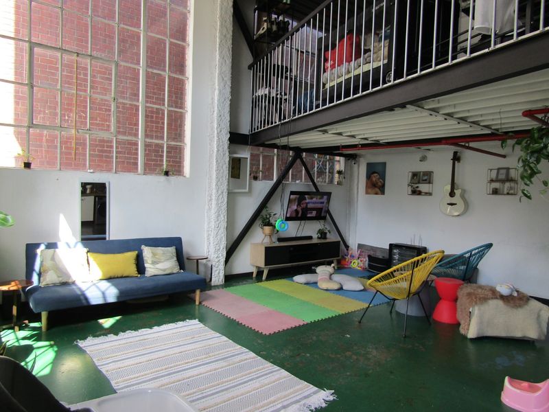 Spacious industrial-style living area featuring high ceilings, large steel-framed windows, exposed brickwork, and a mezzanine level with metal railing.