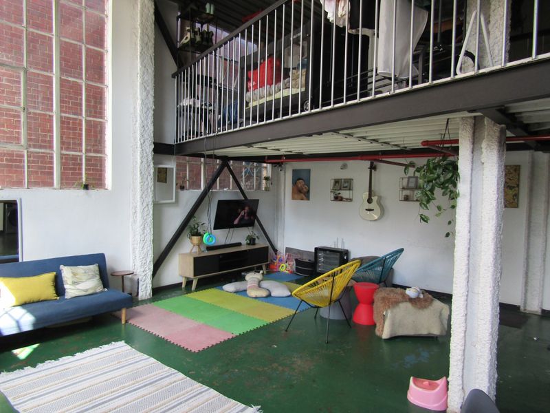 Spacious industrial-style living area featuring a mezzanine level, high ceilings with exposed pipes, large windows, and original brickwork elements.