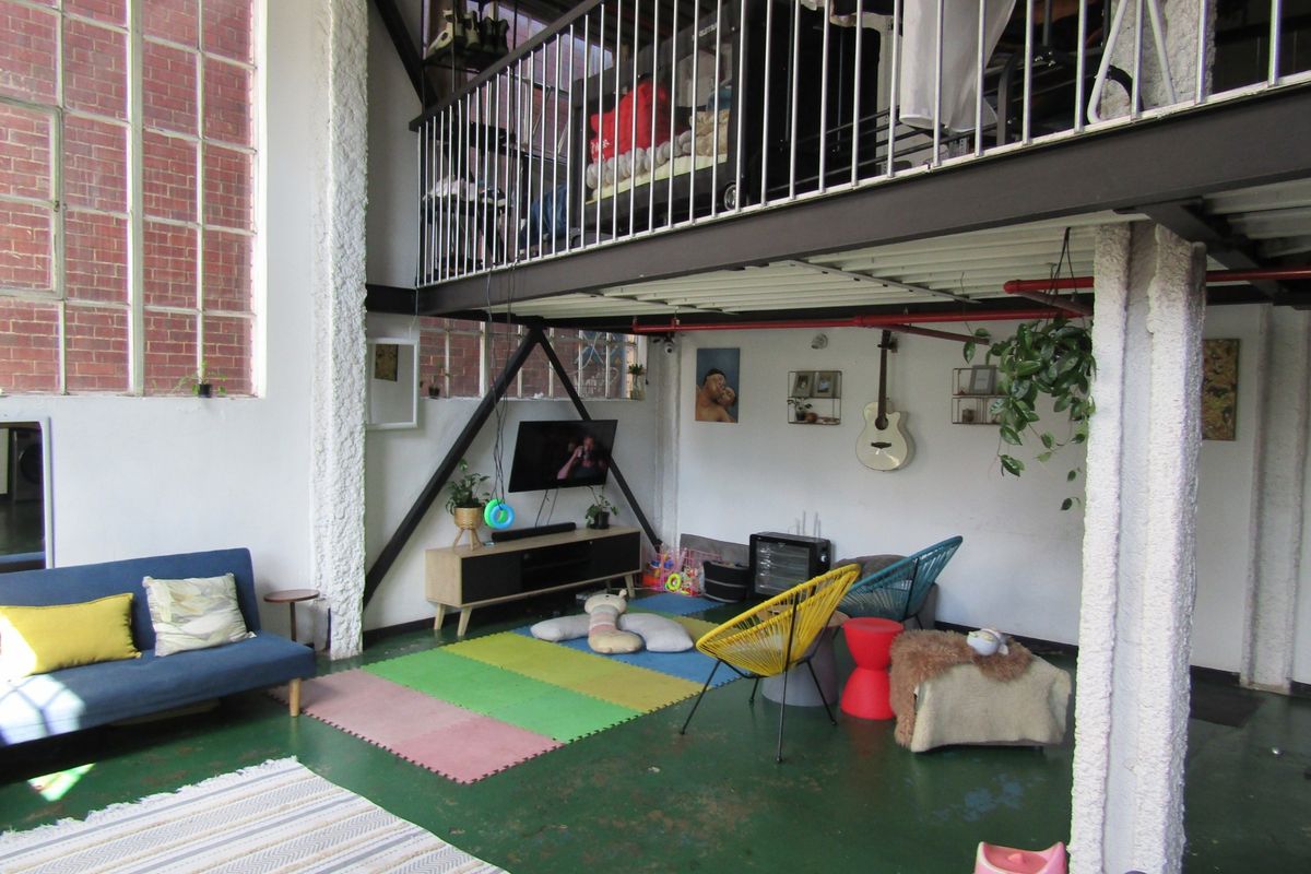Spacious industrial-style living area featuring a mezzanine level, high ceilings with exposed pipes, large windows, and original brickwork elements.