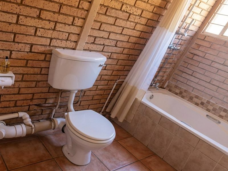 A functional bathroom featuring exposed brick walls, ceramic floor tiling, a built-in bathtub with shower mixer, and a standard toilet.