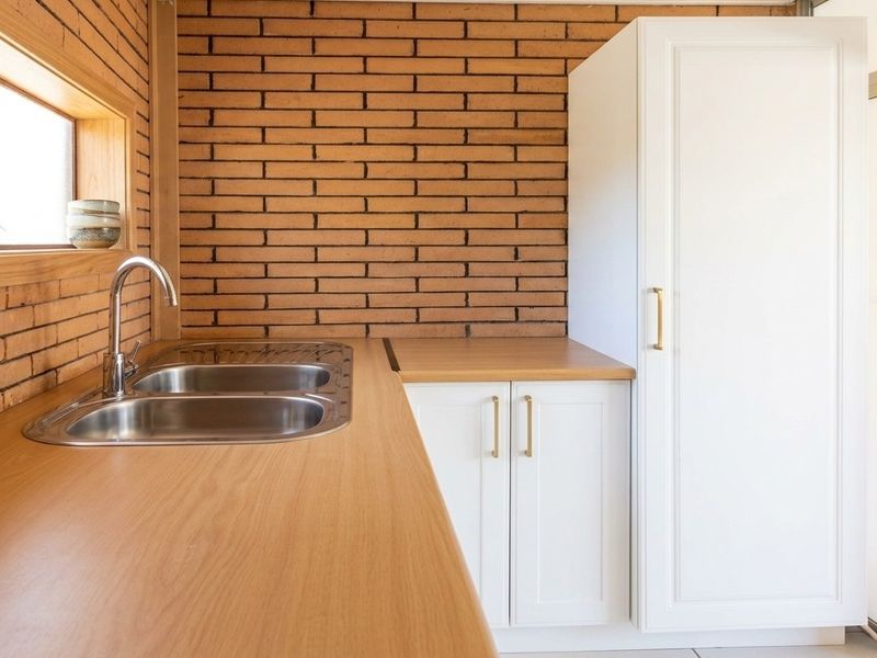 A clean, modern kitchen area featuring exposed brick walls, white cabinetry, timber countertops, and a stainless steel double sink.
