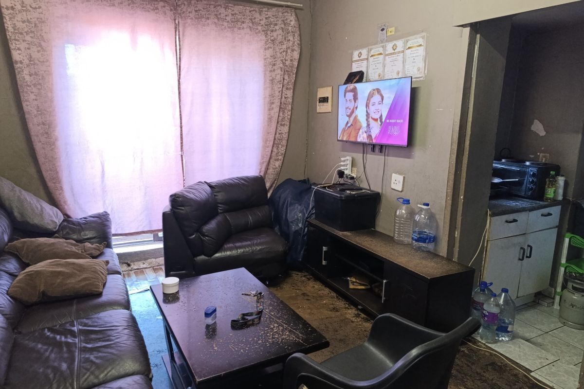A living area with adjacent kitchen space, featuring tiled floors and wall-mounted television, requiring cosmetic updates and maintenance to improve the overall condition.
