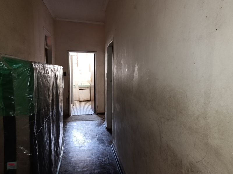 A narrow hallway featuring original parquet flooring, leading towards a bathroom area. The space requires cosmetic updates to the wall finishes.