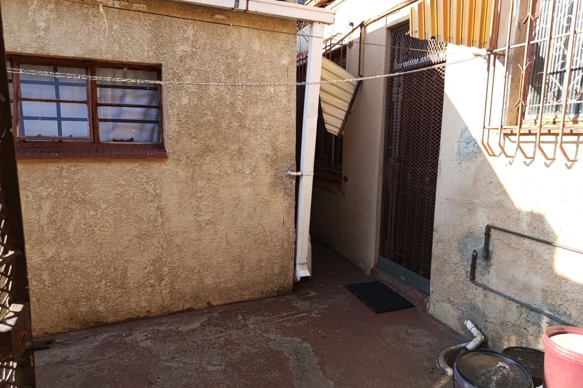 A narrow rear exterior passage featuring concrete paving, security bars on windows, and external plumbing fixtures, showing signs of significant wear and maintenance needs.