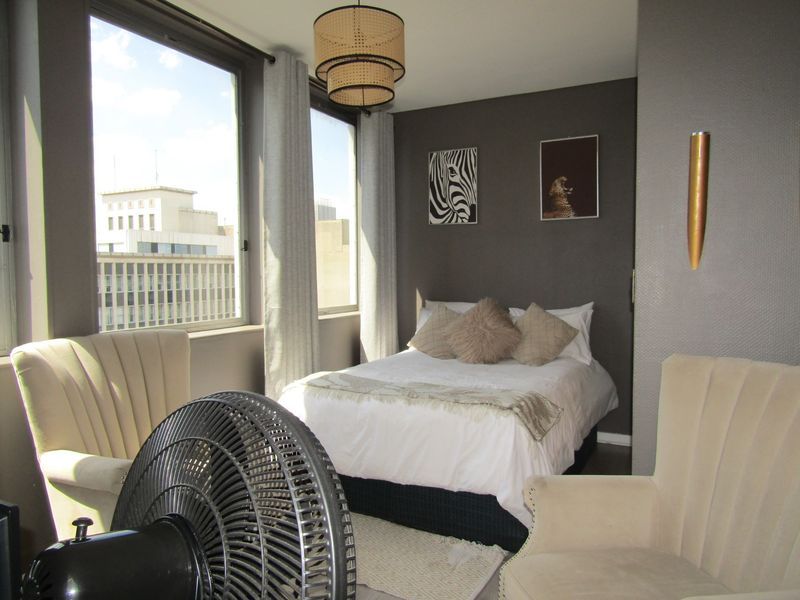 A well-lit bedroom featuring large windows with city views, dark accent wall, and modern wall-mounted lighting.
