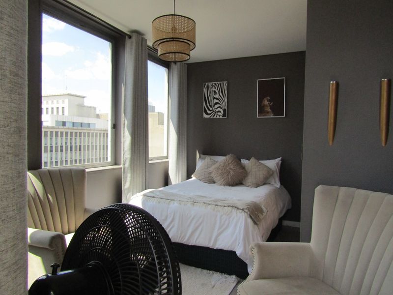 A well-lit bedroom featuring large windows with city views, dark textured accent walls, and modern wall-mounted lighting fixtures.