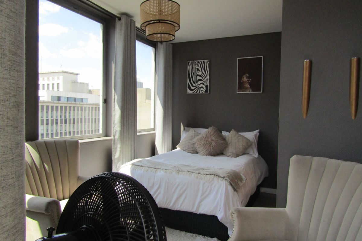 A well-lit bedroom featuring large windows with city views, dark textured accent walls, and modern wall-mounted lighting fixtures.