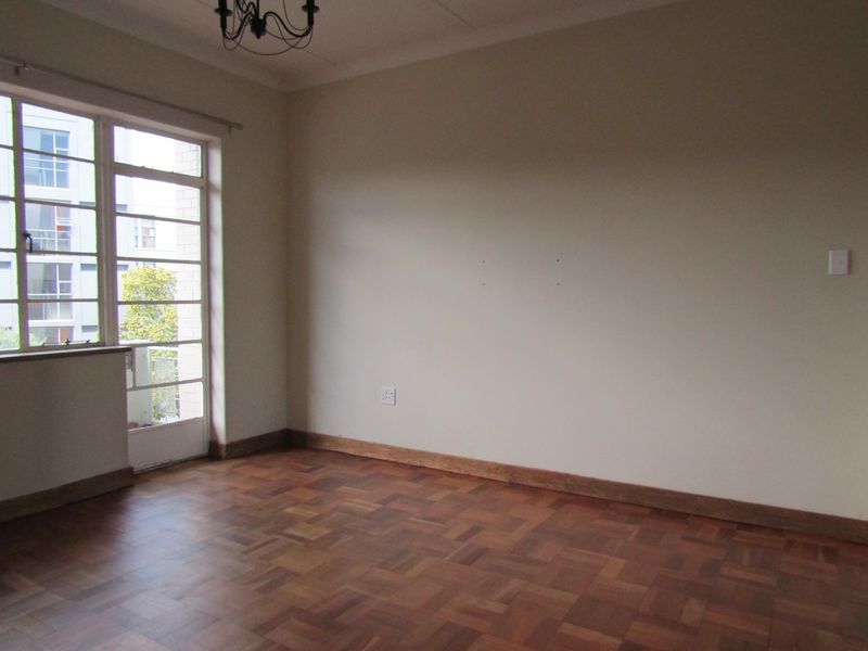 A bright, empty living room featuring classic parquet flooring, timber skirting boards, and a steel-framed glass door leading to an outdoor area.