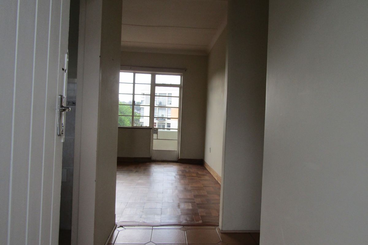 An empty living area featuring parquet flooring, tiled transition space, and large metal-framed windows providing natural light and access to an exterior balcony.