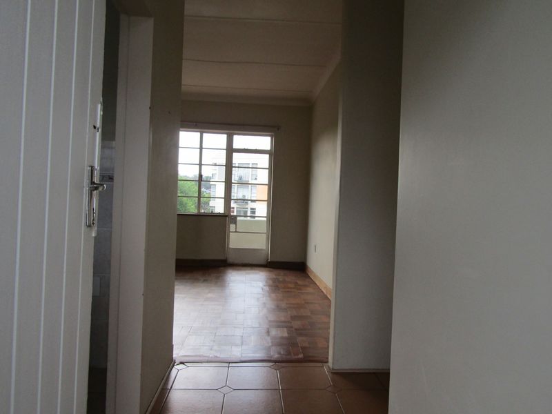 An empty living area featuring parquet flooring, tiled transition space, and large metal-framed windows providing natural light and access to an exterior balcony.