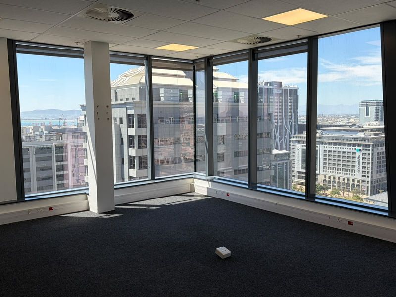 A bright, empty corner office space featuring floor-to-ceiling windows with expansive city views, carpeted flooring, and a modern suspended ceiling.