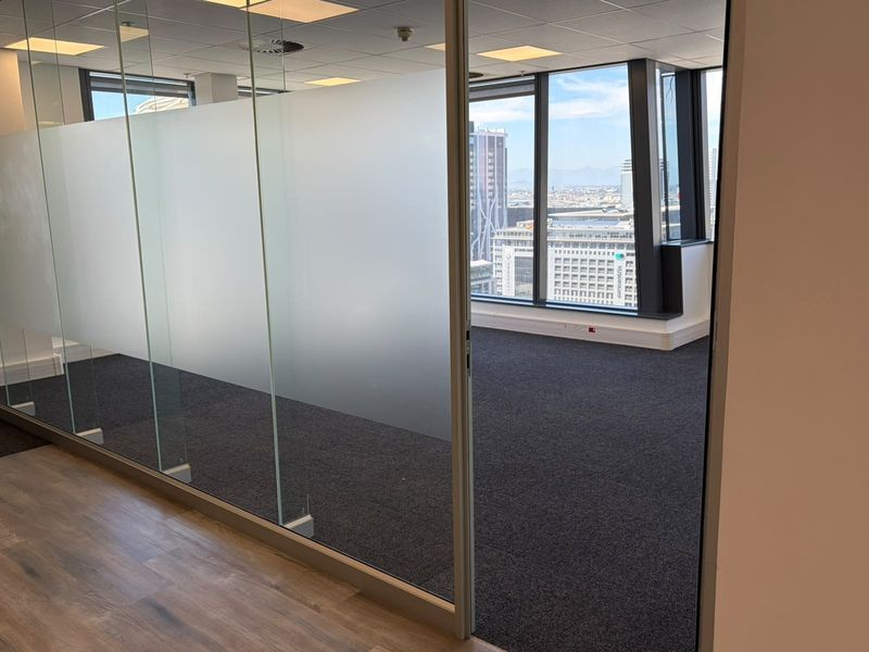 Modern office space featuring glass partitions, carpeted flooring, and floor-to-ceiling windows offering expansive city views.